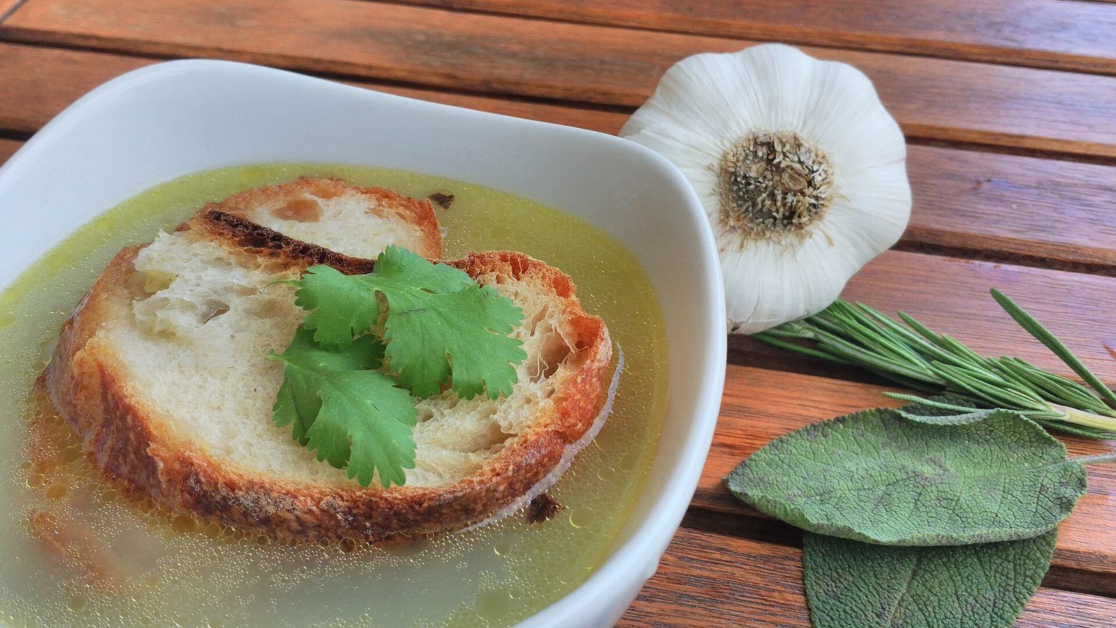 Bol de bouillon à l'ail avec une tranche de pain grillé, à côté d'une tête d'ail, de feuilles de sauge et de romarin