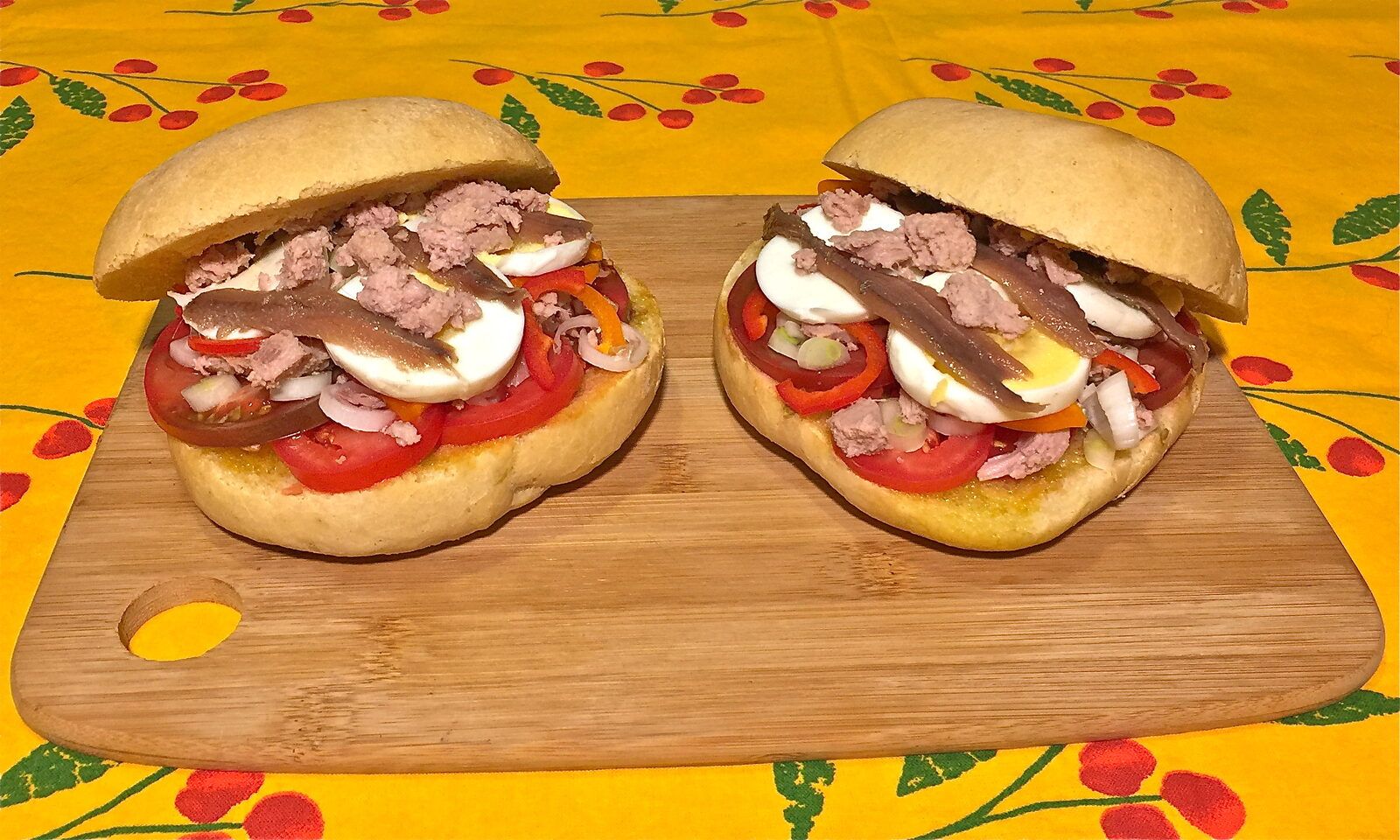 Deux pans bagnats ouverts sur une planche en bois, chaque petit pain rond garni de tranches de tomate, d’œuf dur, de thon émietté et de filets d’anchois