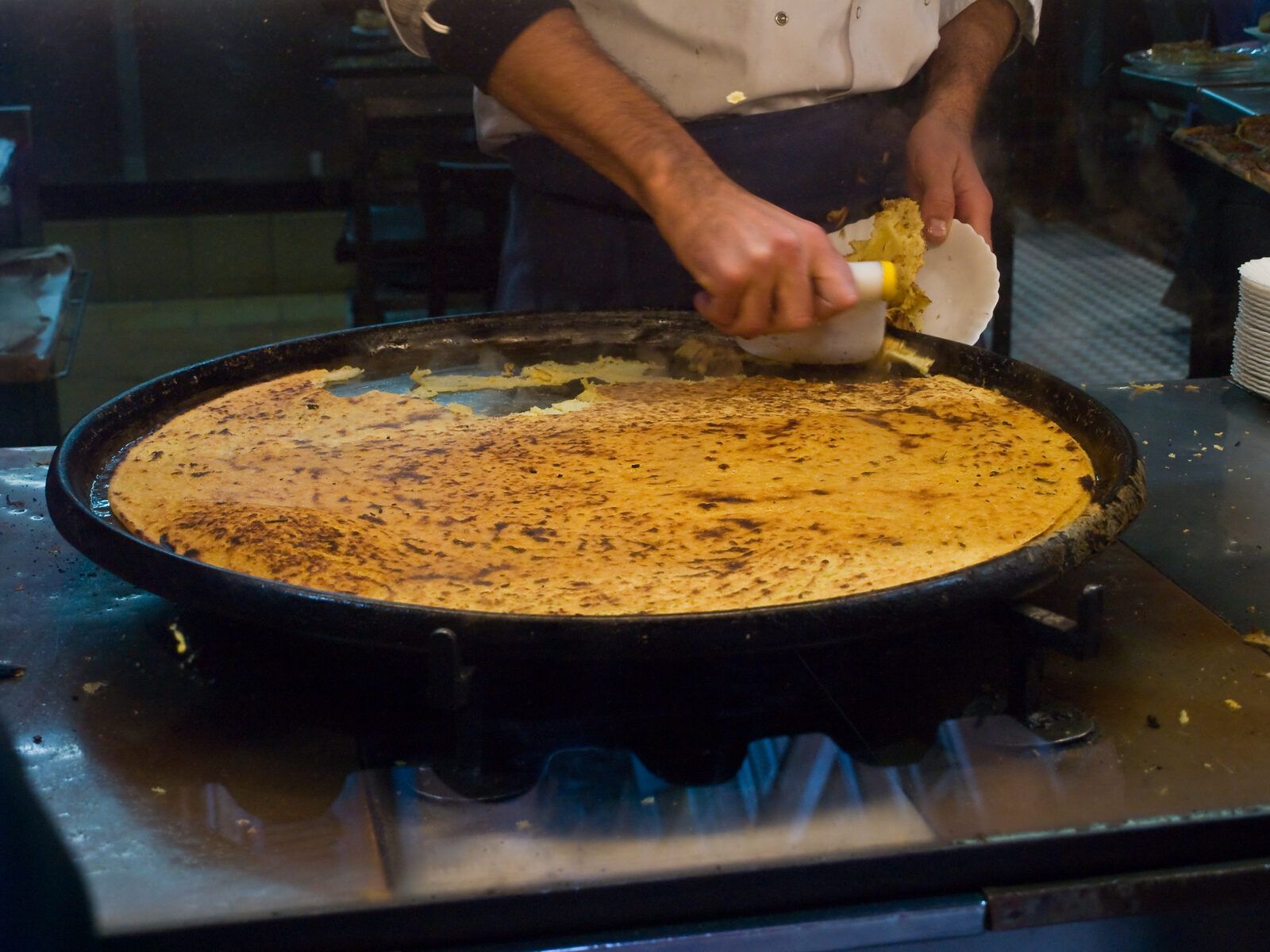 A cook scraping hot socca off its large round plate, just out of the oven in the old town of Nice